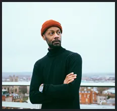A man wearing a red beanie and black turtleneck standing outdoors with arms crossed, city buildings visible in the background under a cloudy sky.