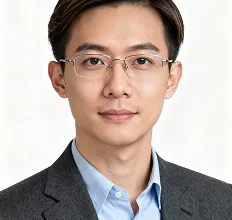 Portrait of a young Asian man wearing glasses, a light blue shirt, and a dark suit jacket, looking directly at the camera with a neutral expression against a plain light background.