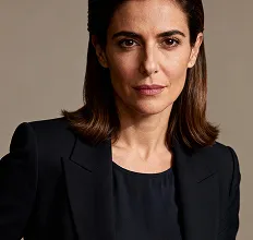 Confident woman with straight dark brown hair wearing a black blazer and black top, posing against a plain brown background.