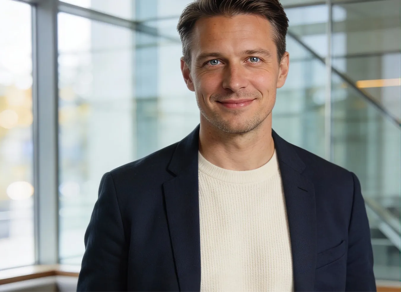 Smiling man with blue eyes wearing a white sweater and dark blazer standing indoors in front of large windows with a blurred background.