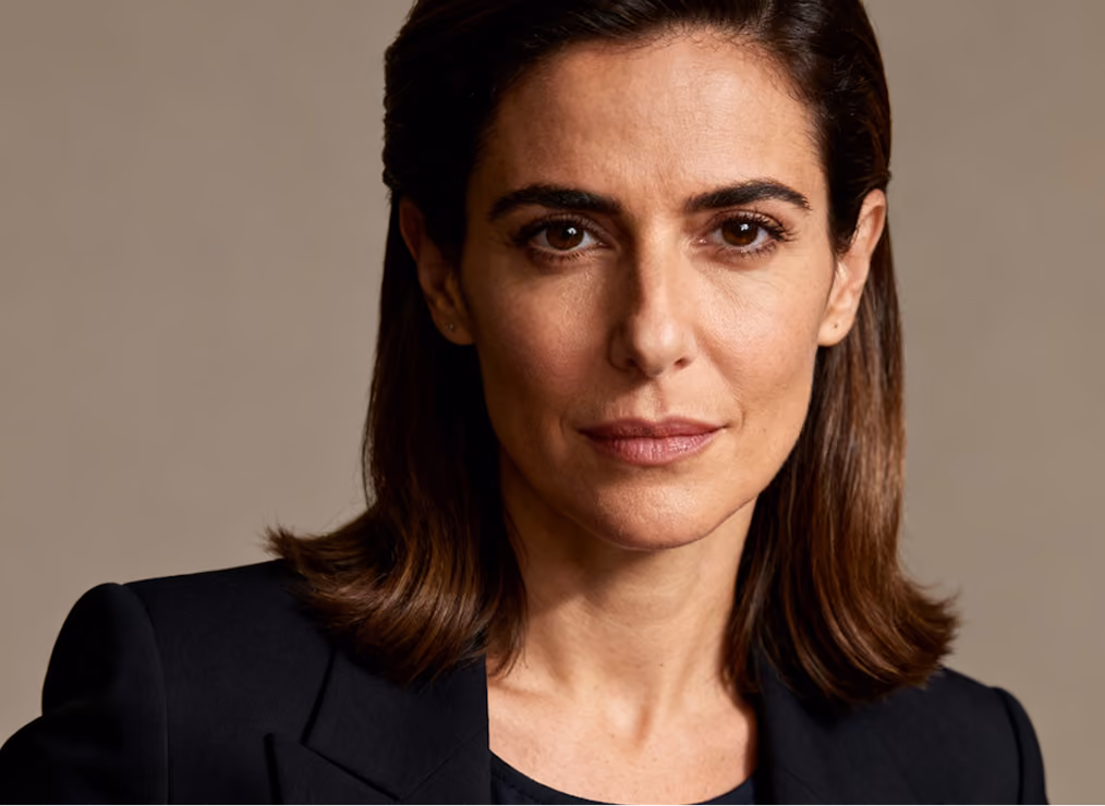 Close-up portrait of a woman with medium-length brown hair wearing a black blazer and looking directly at the camera against a plain beige background.