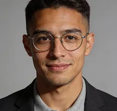 Portrait of a young man with short dark hair wearing round eyeglasses, a gray shirt, and a black blazer, smiling slightly against a plain gray background.