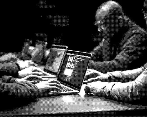 Four people sitting at a wooden table working on laptops, with one man's focused face visible, in a dimly lit room.
